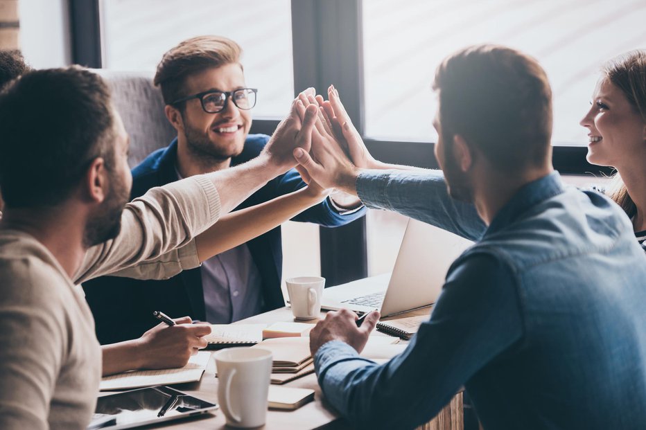 Fotografija: Cheerful young people giving each other high-five with smile while sitting at the office table on business meeting