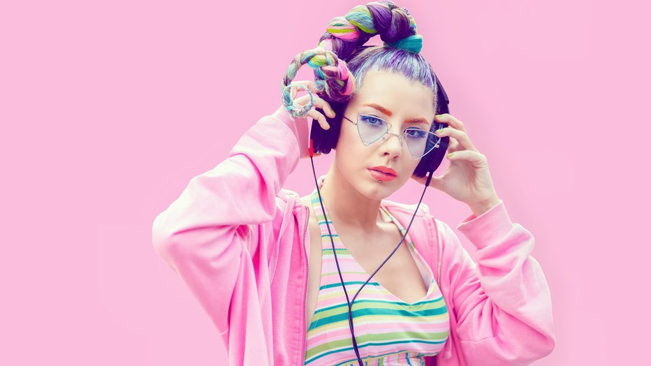 Fotografija: Confident fashionable funky young woman with creative hairstyle wearing cool hoodie standing against pink background and listening music on big headphones while looking at camera