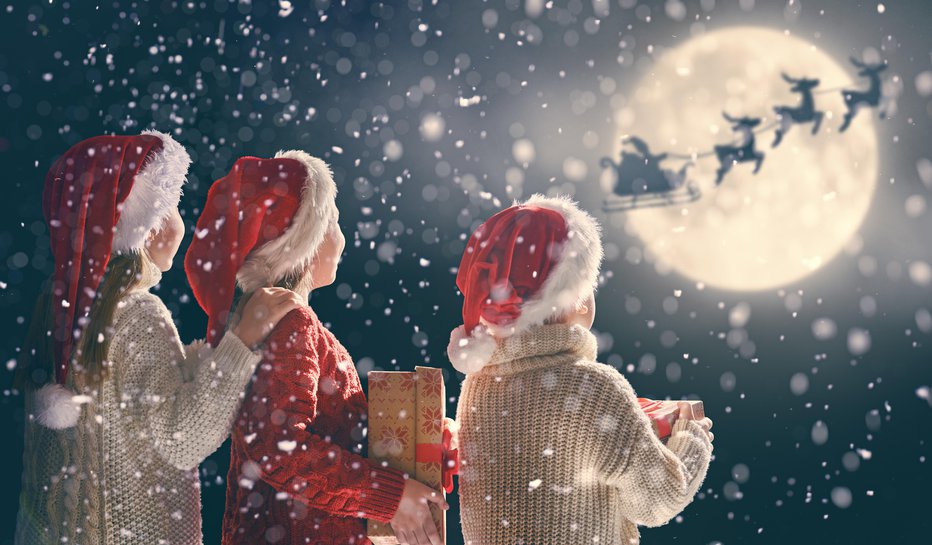 Fotografija: Merry Christmas and happy holidays! Cute little children with xmas presents. Santa Claus flying in his sleigh against moon sky. Kids enjoying the holiday with gifts on dark background.
