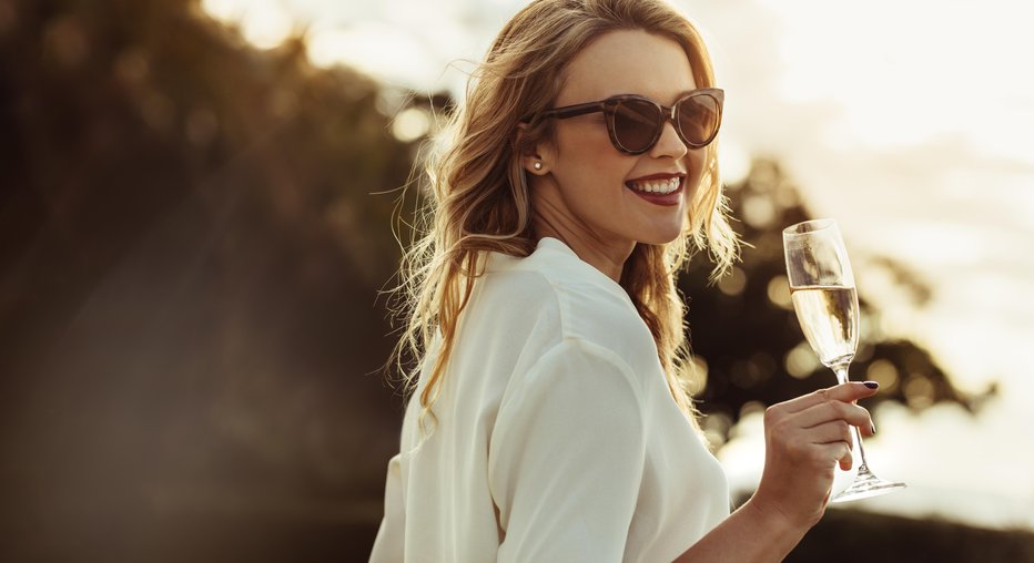 Fotografija: Elegant woman in sunglasses with a glass of wine outdoors. Smiling caucasian female having wine and looking backwards.